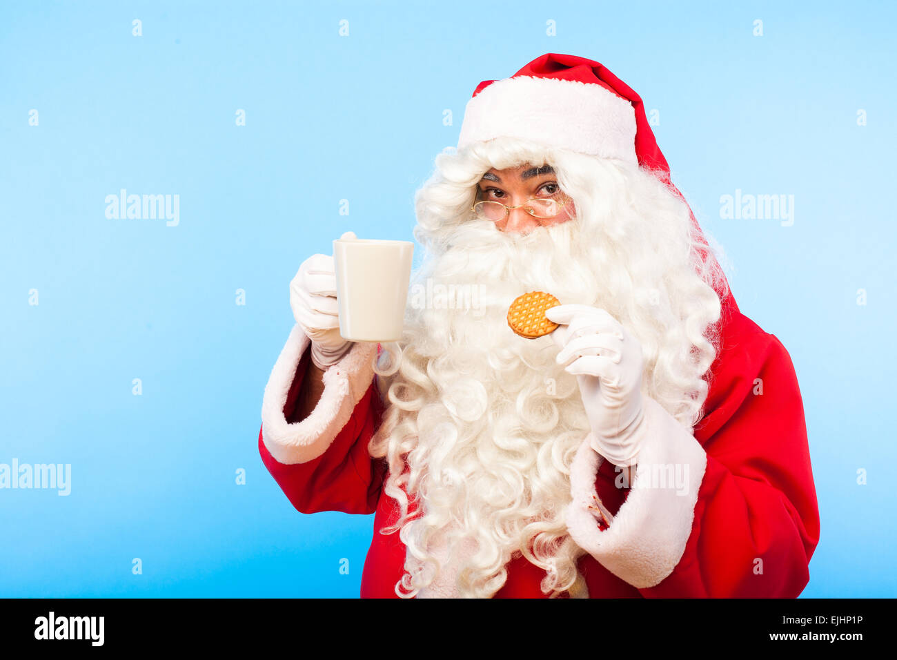 happy santa looking at camera with a cup of coffee and biscuit in hand, on blue background Stock Photo