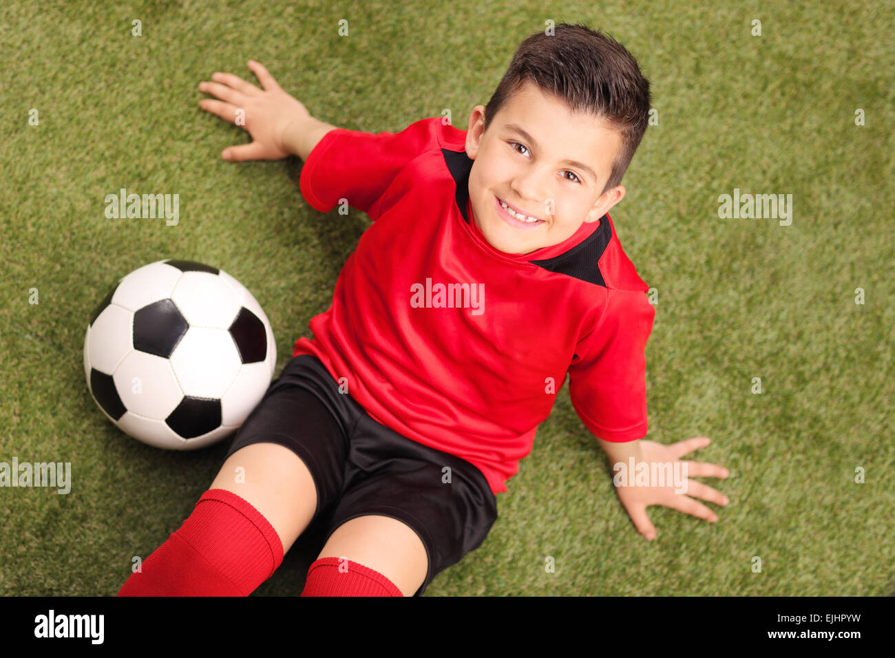 High angle shot of a junior soccer player in red jersey, sitting on a green field and looking at the camera, with a soccer ball Stock Photo
