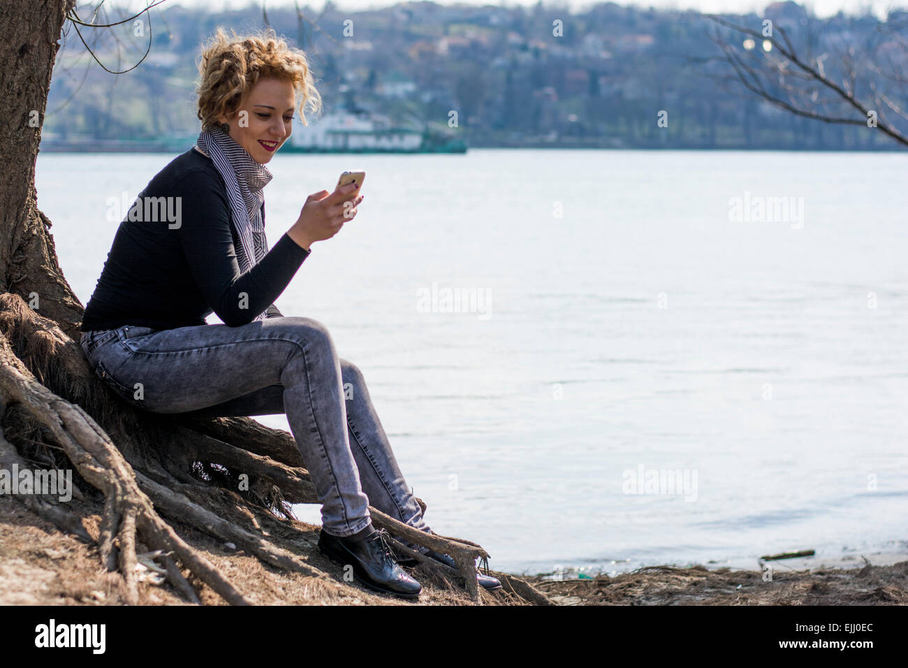 Pretty young woman with blond curly hair typing on the phone sitting on the root of the big tree near the river.She is smiling a Stock Photo