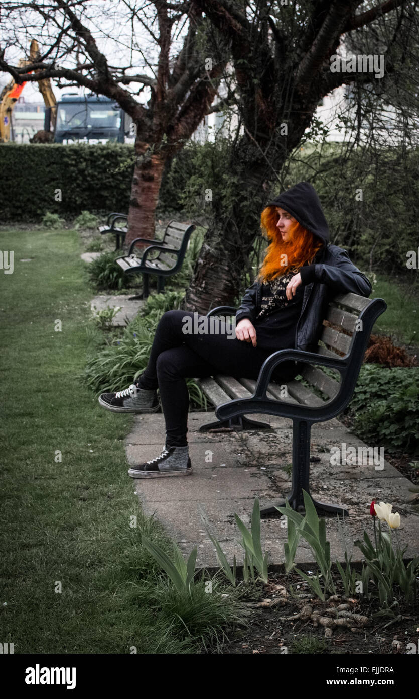 Woman with bright orange hair sat on a park bench in a city park in Hampshire, UK with hood up with tulips in the foreground. Stock Photo