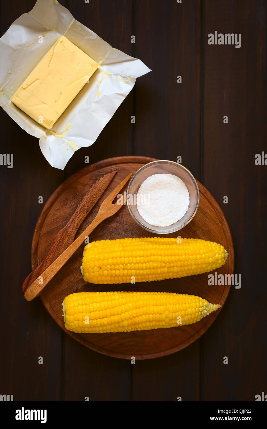 Overhead shot of two cobs of cooked sweet corn on wooden plate with salt and cutlery, with a piece of butter above Stock Photo