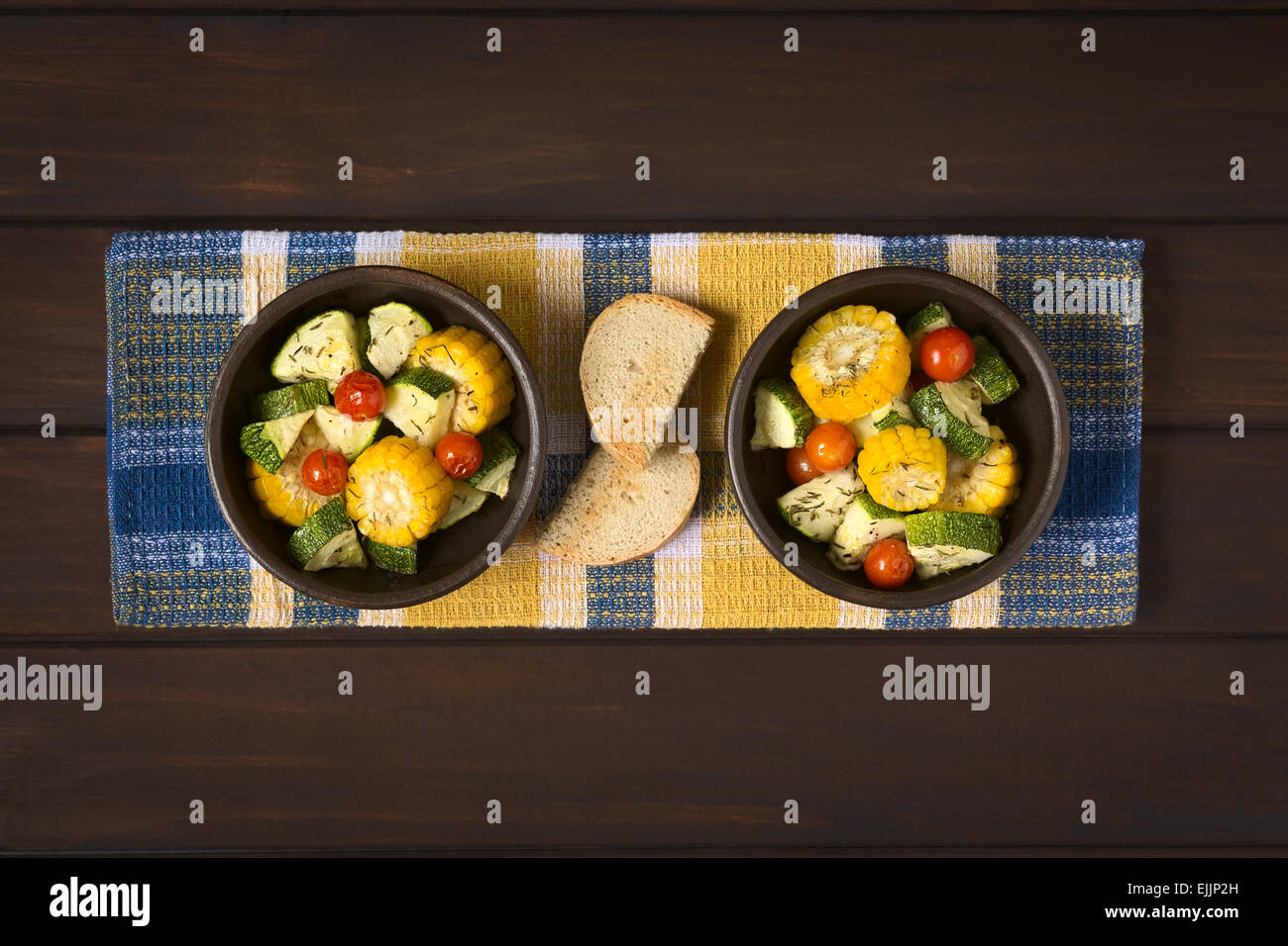 Overhead shot of two bowls of baked vegetables of sweet corn, zucchini, cherry tomato with thyme, toasted bread slices beside Stock Photo