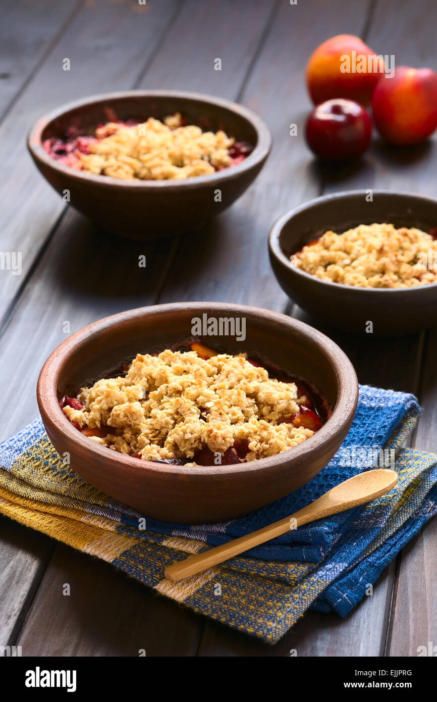 Three rustic bowls filled with baked plum and nectarine crumble or crisp, photographed on dark