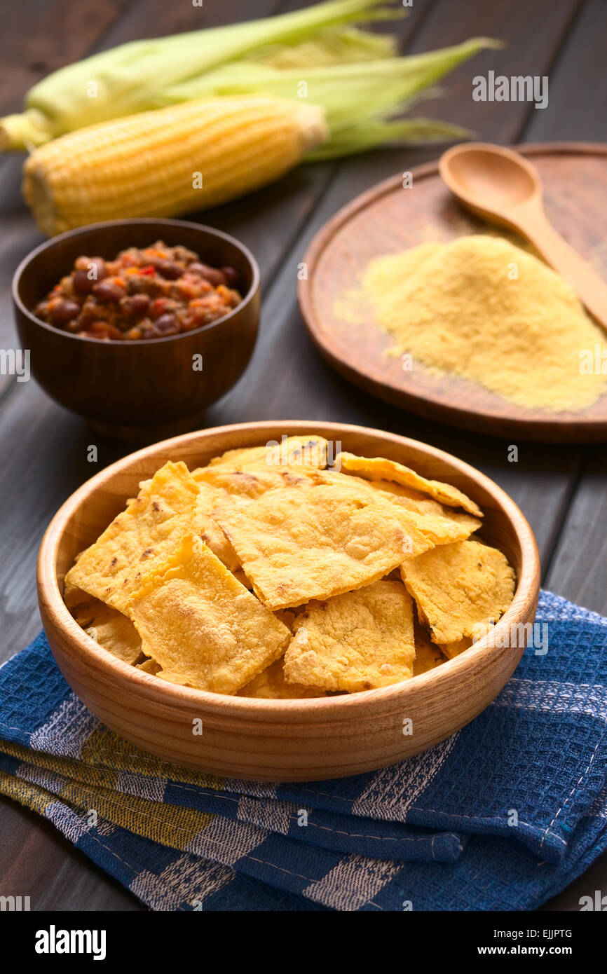 Wooden bowl of homemade baked corn chips with cobs of corn, cornmeal and a bowl of chili con carne in the back Stock Photo