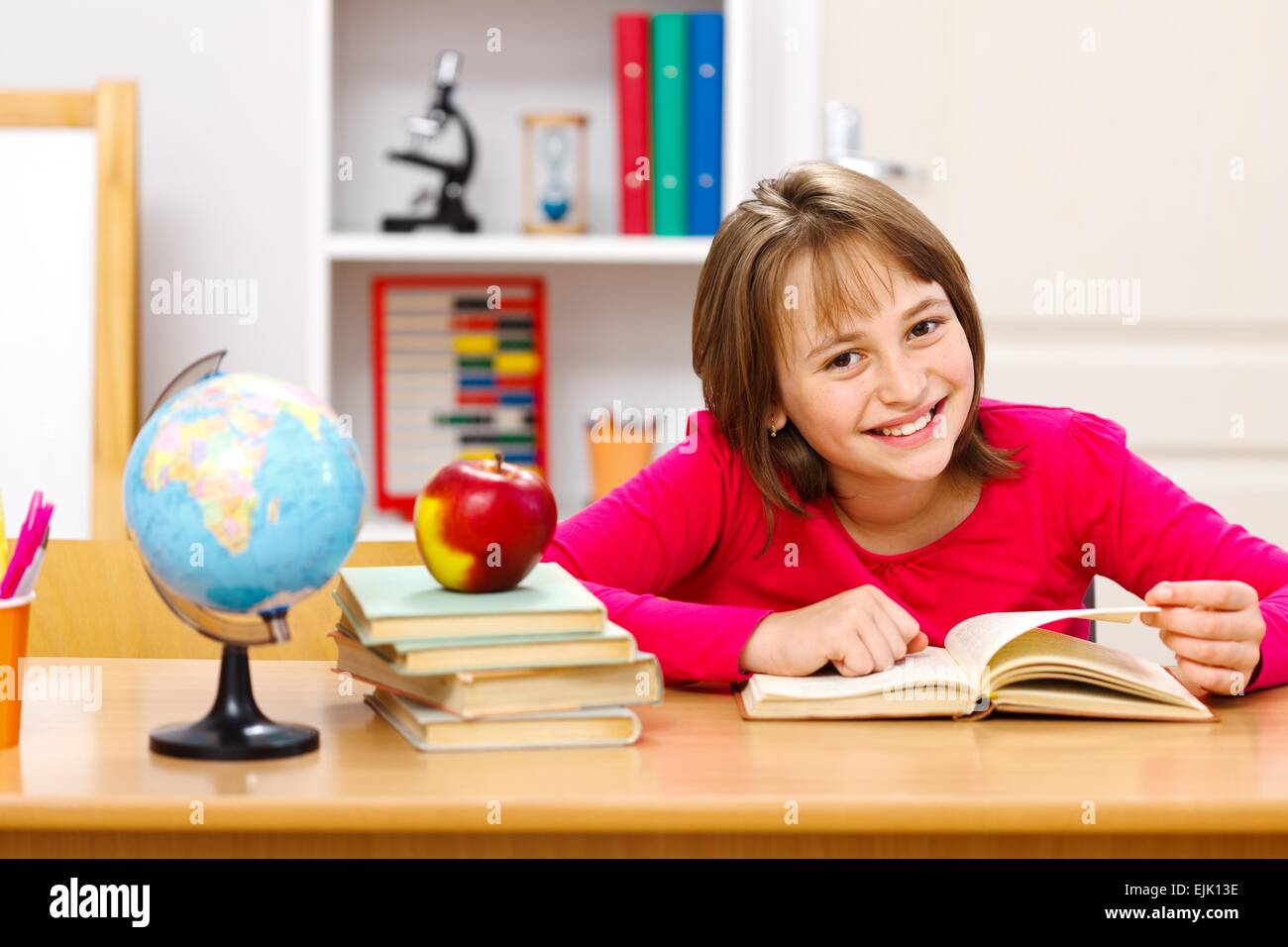 Pretty schoolgirl reading in classroom. Earth globe and whole apple on other stacked books Stock Photo