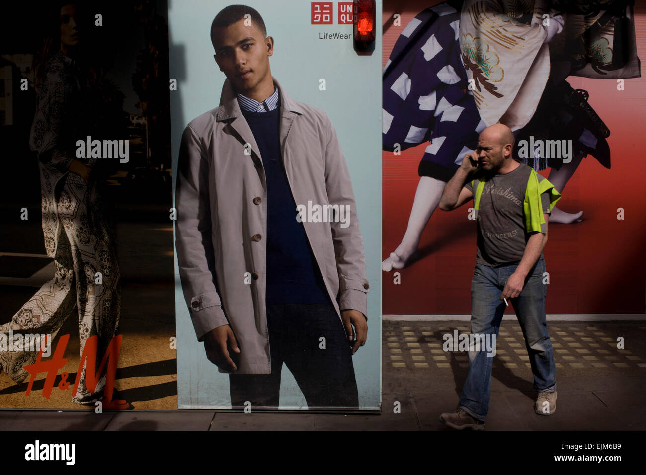 A workman talking on his smartphone walks past a H&M and Uniqlo menswear  poster outside central London shop. The models on the poster are perfect  ideals, models of perfection while the person