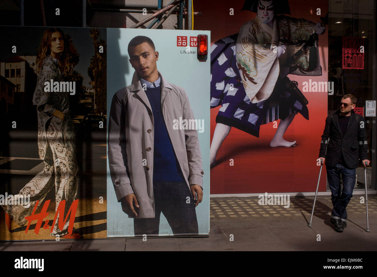 Man with walking crutches in front of H&M and a Uniqlo menswear poster  outside central London shop. The injured man waits for a bus but is  surrounded by fashion imagery with the