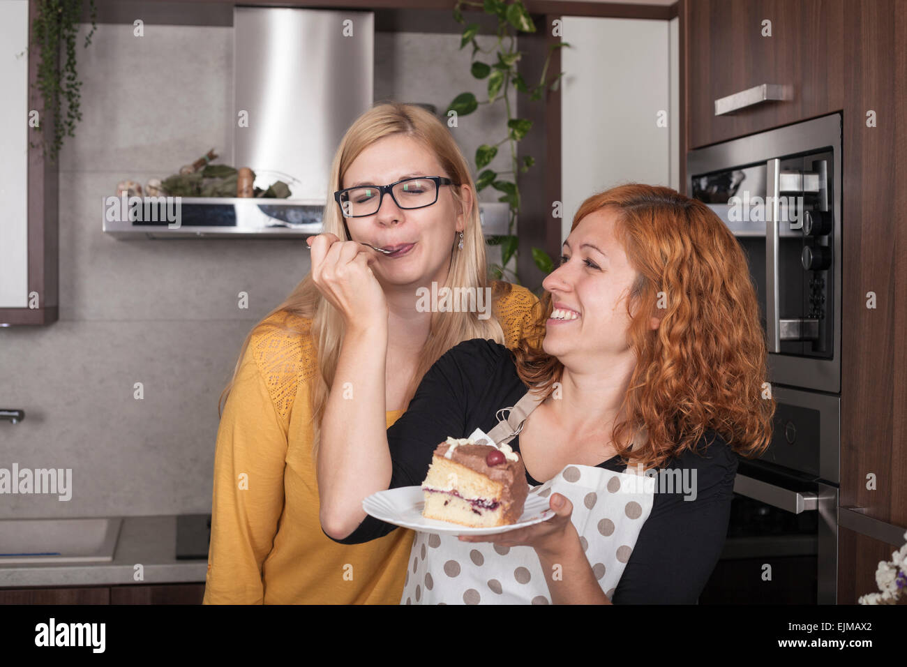 Delighted girlfriends enjoying cake and feeding each other in the kitchen at home. Stock Photo