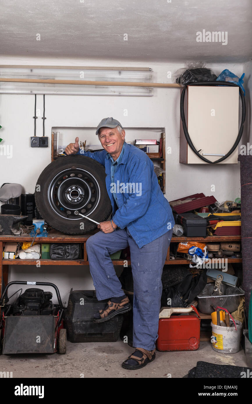 Portrait of happy senior worker with a tire and tool showing thumb up in the garage. Stock Photo