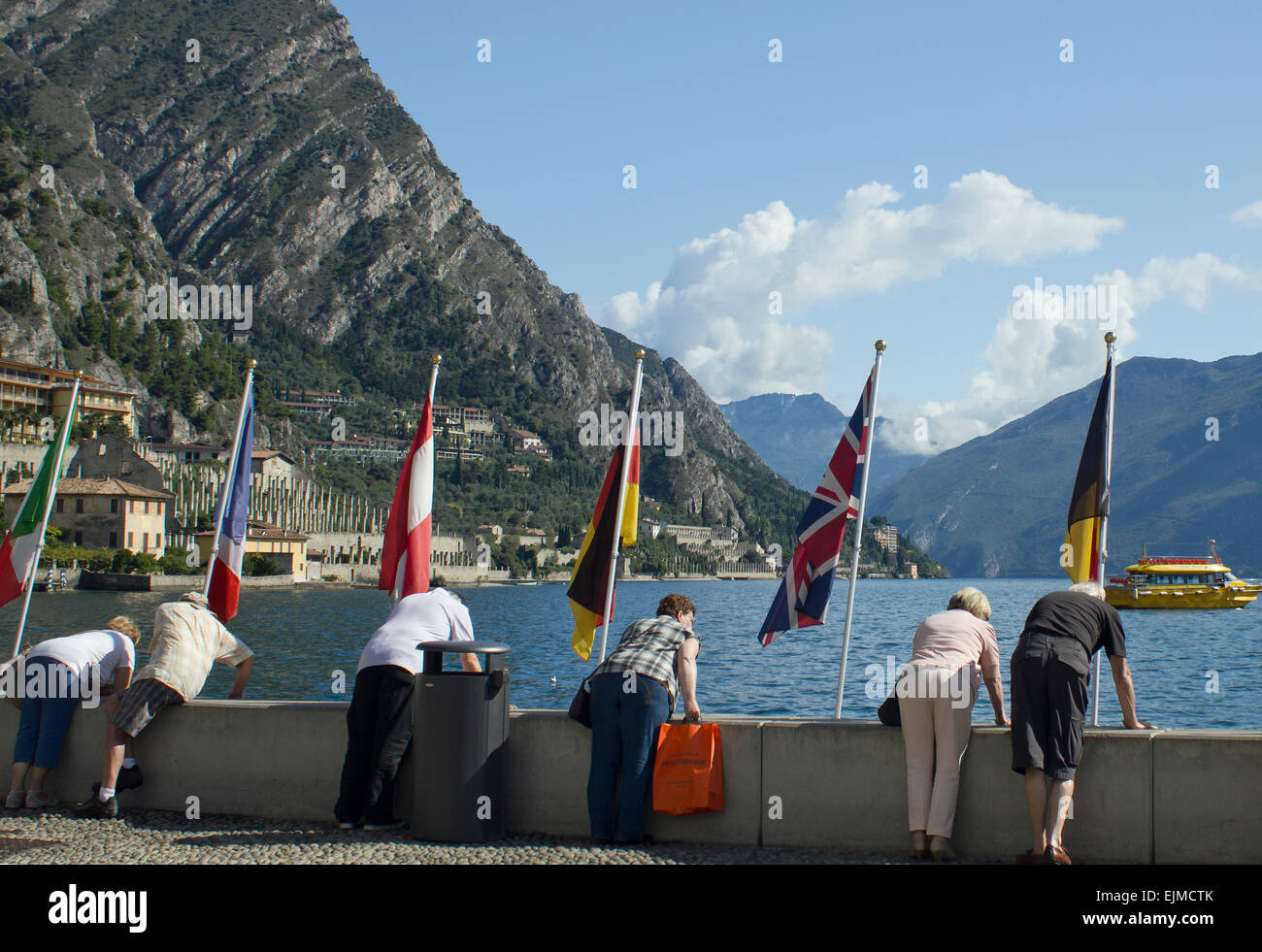 Tourists at Limone,Lake Garda, Italy, bending over a wall to view the lake, with flags of European countries on display. Stock Photo
