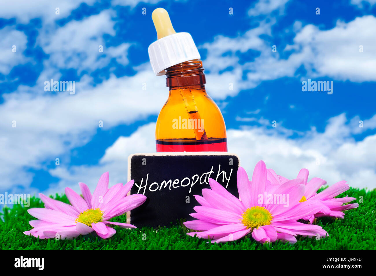 a dropper bottle and some purple flowers on the grass with a blackboard label with the word homeopathy written on it Stock Photo