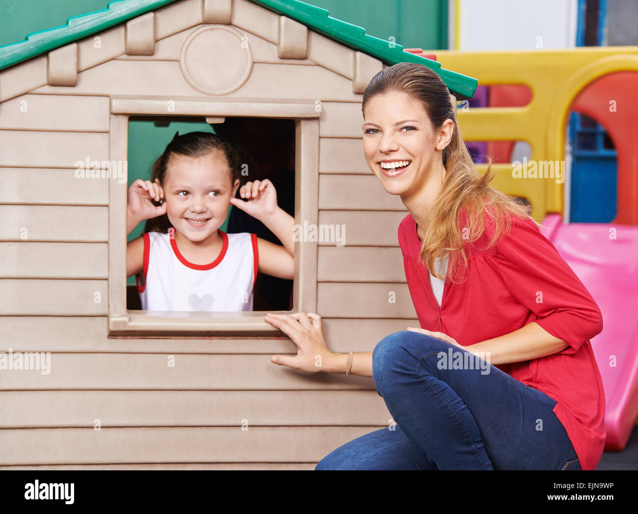 Happy educator playing with girl in playhouse in kindergarten Stock