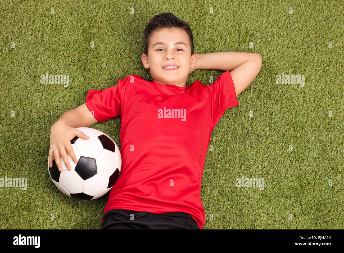 Relaxed youngster in a red football jersey lying on a pitch, holding a football in his hand and looking at the camera Stock Photo