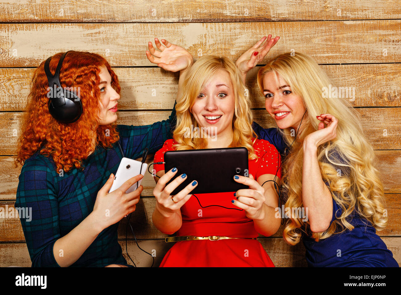Three sisters, blond and red, listening to music on headphones. Girl doing funny ears hands sister, looking at screen Stock Photo