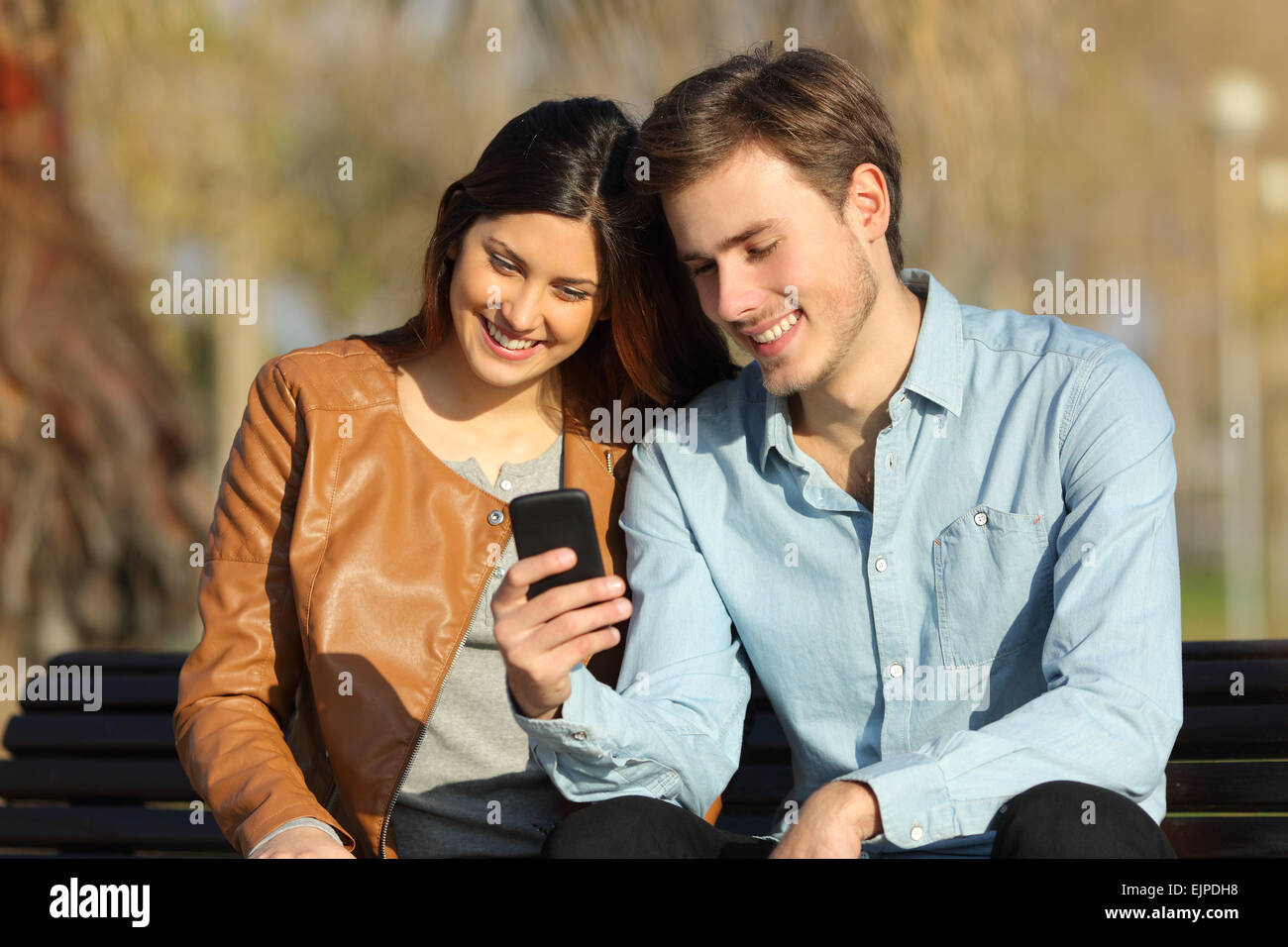 Happy couple watching a smart phone sitting on a bench in a park Stock ...