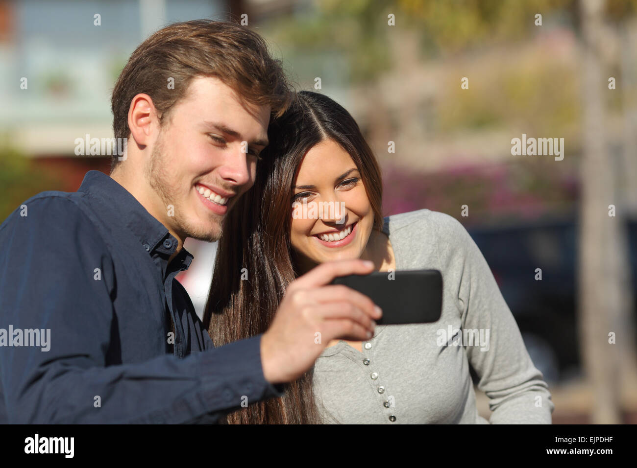 Happy couple watching media in a smart phone outdoors with an urban background Stock Photo
