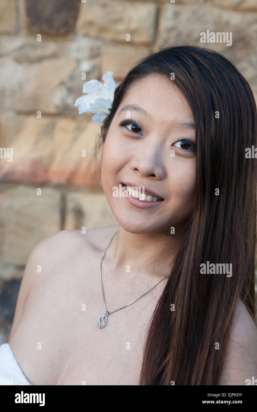 Pretty Asian woman outdoors by a stone wall, in a white dress and with a white flower in her hair Stock Photo