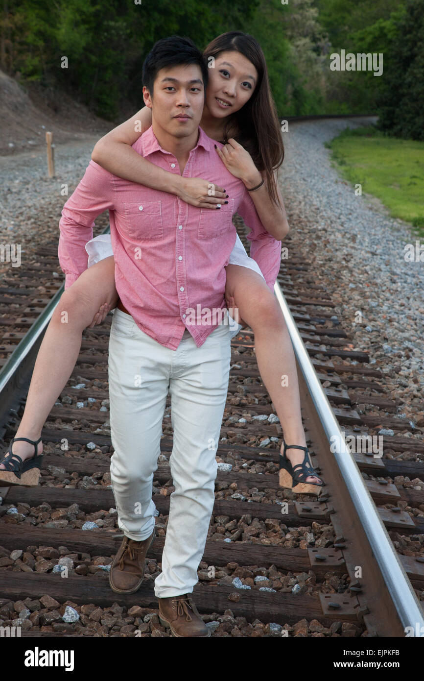 Romantic Asian couple playing outside on railroad tracks, she is riding on his back in a "piggyback" fashion Stock Photo