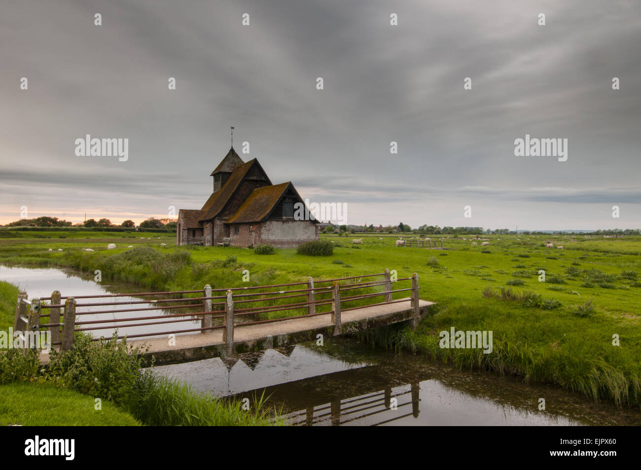View of sheep on grazing marsh with footbridge across flooded ditch and church in evening, St. Thomas a Becket Church, Fairfield, Walland Marsh, Romney Marsh, Kent, England, May Stock Photo