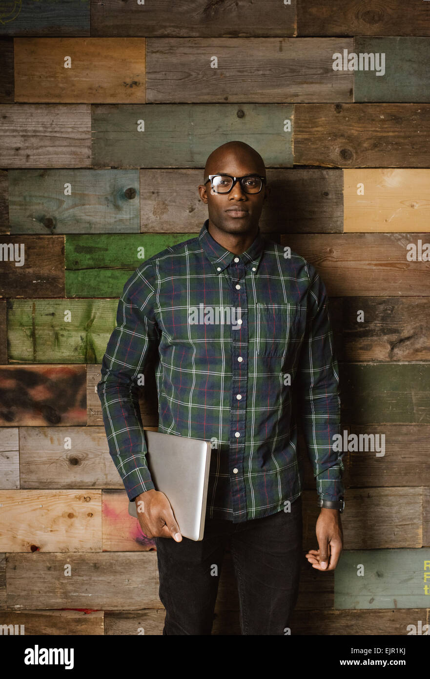 Portrait of serious young african man with a laptop looking at camera while standing against a wooden wall in office. Stock Photo