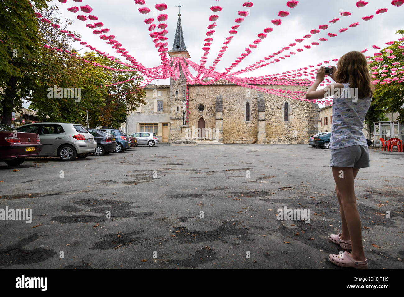 Teenage girl takes a photograph of a display of pink and white paper flowers draped across a town square Villefranche de lonchat Stock Photo