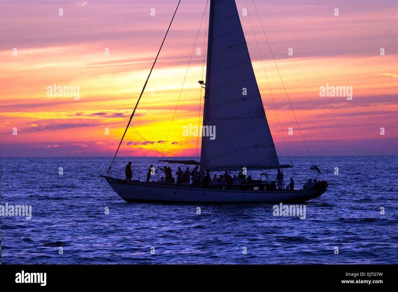 A Sailboat full of party people on vacation sails along the ocean as the sun sets in the background Stock Photo