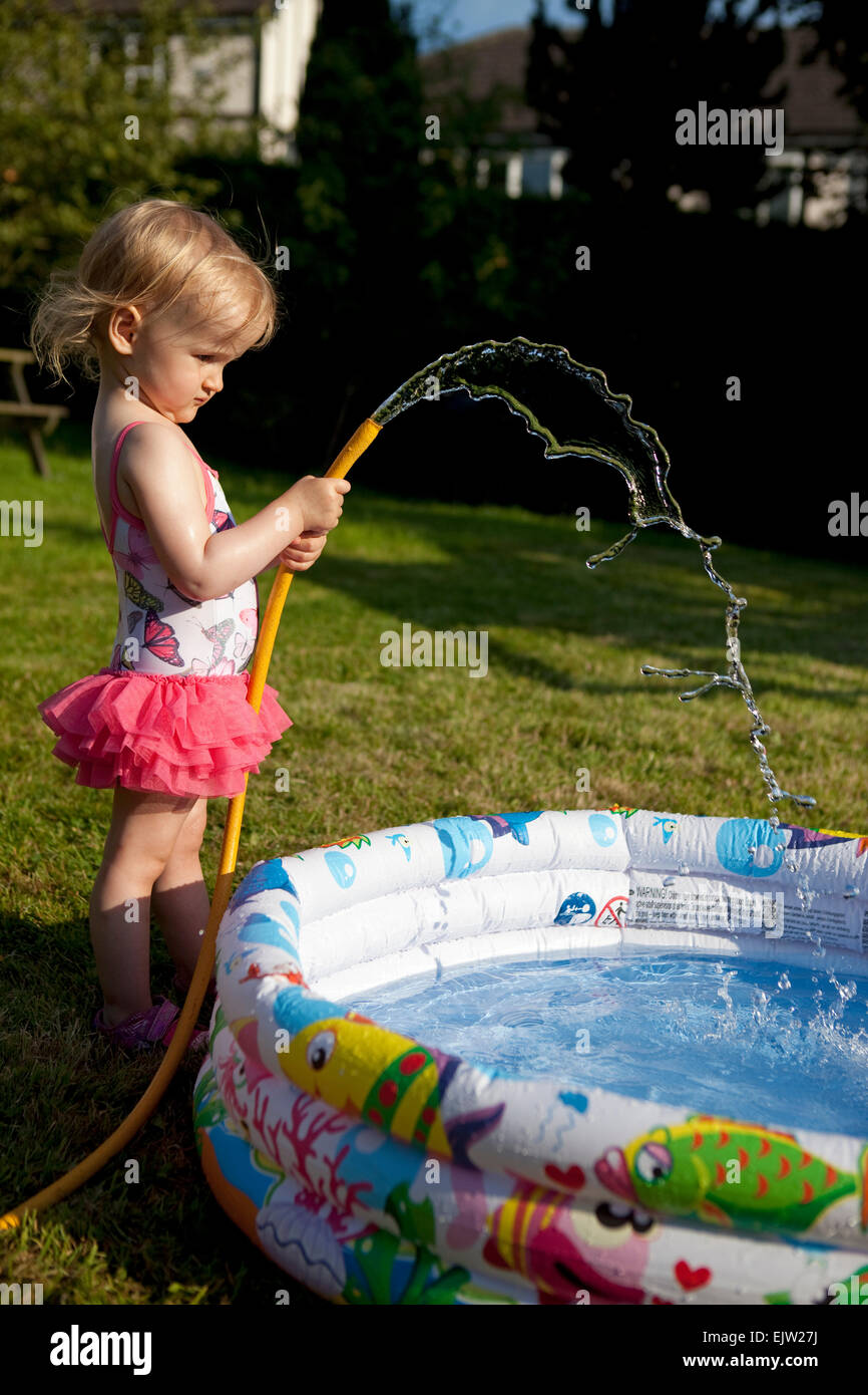 Young girl filling a paddling pool with water from a hosepipe in summer