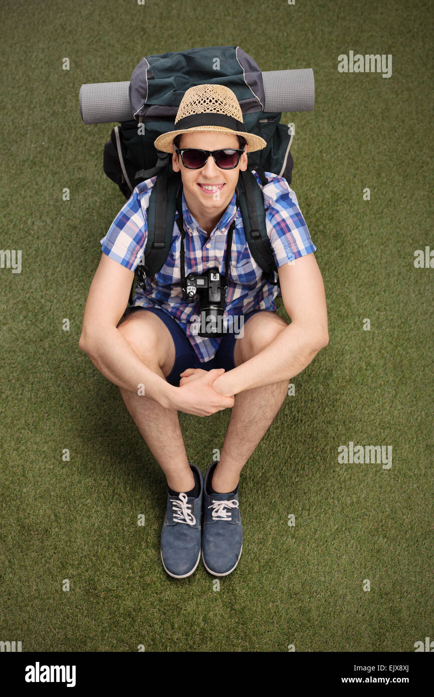 Vertical shot of a cheerful young hiker sitting on grass, carrying a backpack with hiking equipment and looking at the camera Stock Photo