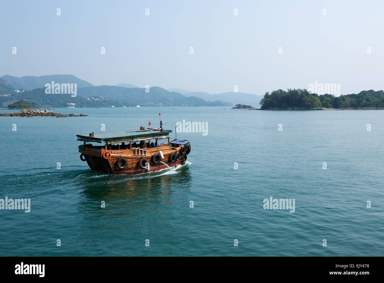 Small Traditional Chinese boat leaving a fishing village in Hong Kong, traveling towards islands across a calm sea leaving a wake on the surface. Stock Photo