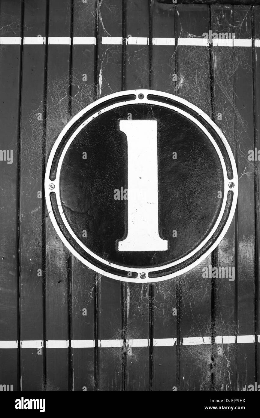 Numeral one, old fashioned sign, on circular cast metal and painted, mounted on wooden paneled wall in black and white retro Stock Photo