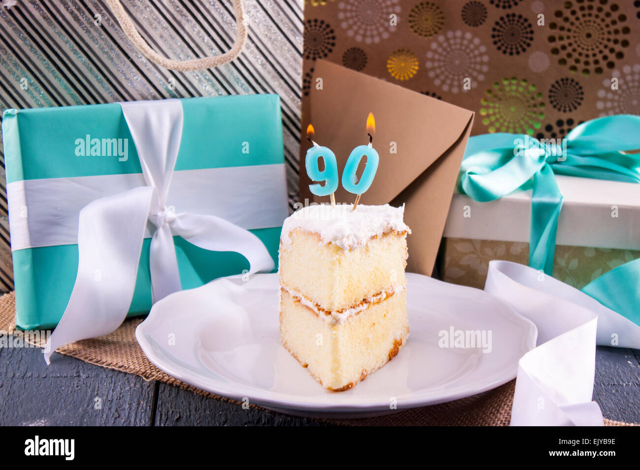 Slice of birthday cake with '90' candle for ninetieth birthday placed with gifts and cards. Stock Photo