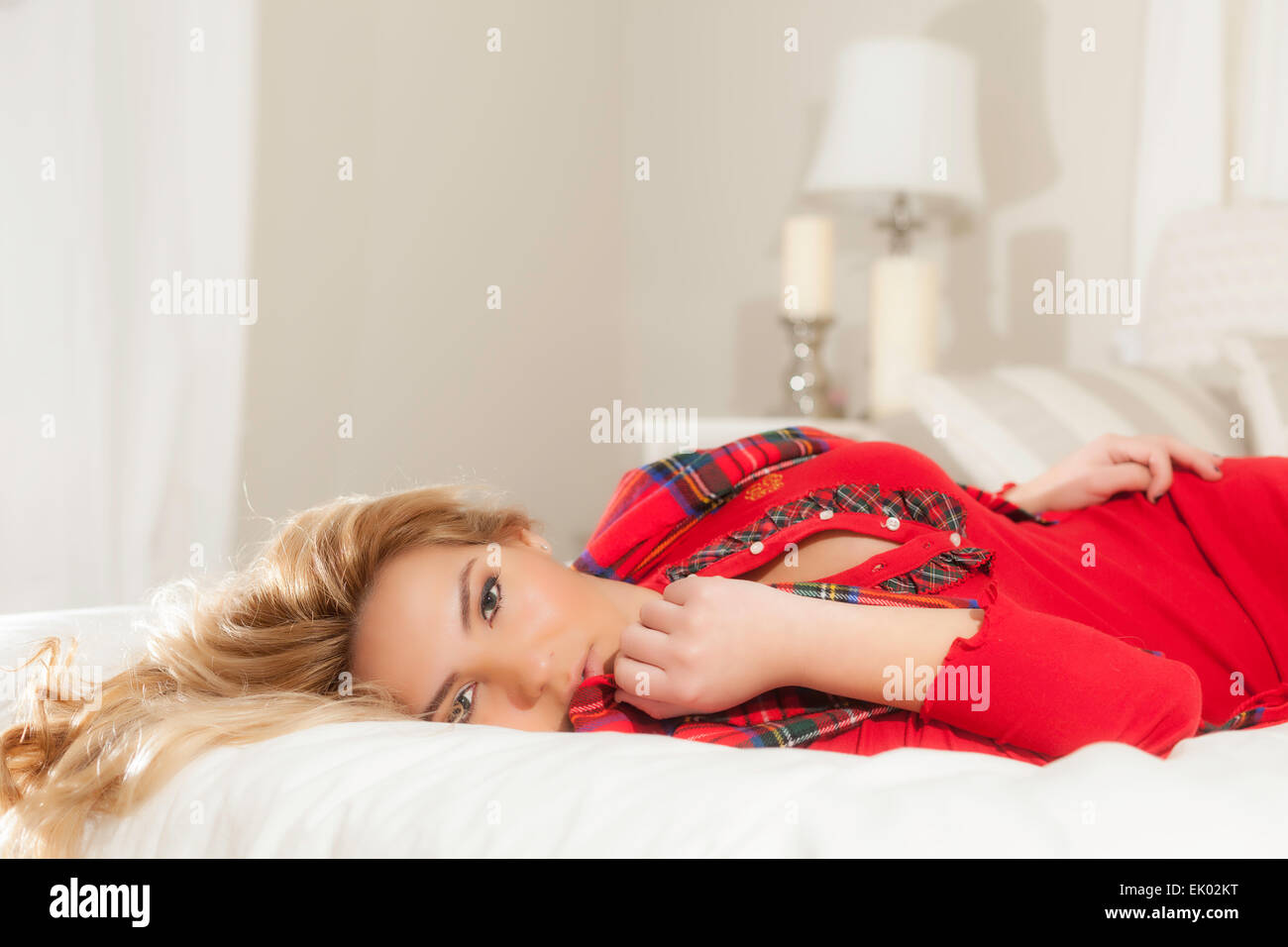 A pretty Latina blonde girl in red Christmas clothes laying on a white bed with a lamp in the background. Stock Photo