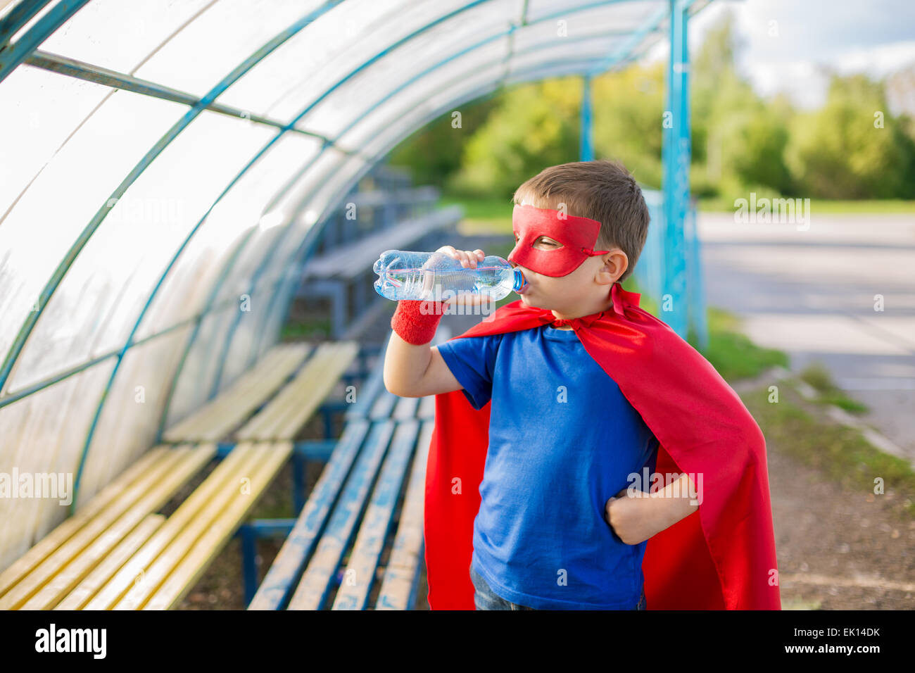 Boy dressed in cape and mask standing under canopy and drinking water from a bottle Stock Photo