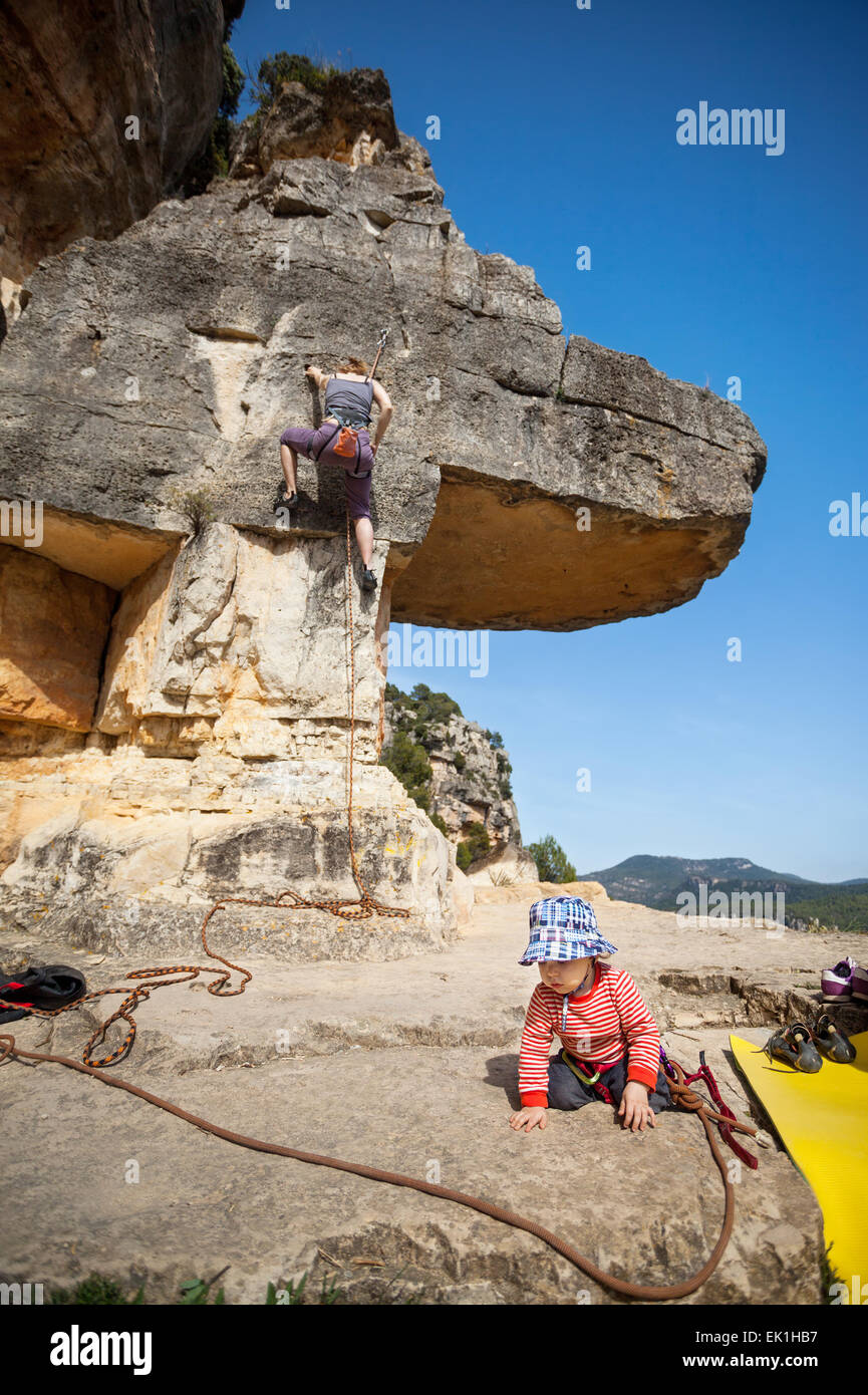 Toddler boy playing at foot of mountain while his mother climbing at the background. The boy is tied in to the rope. Stock Photo