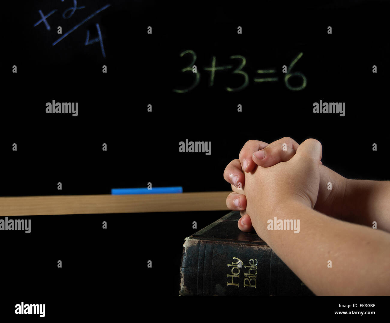 Child's hands folded in prayer on Bible in school with chalkboard. Stock Photo