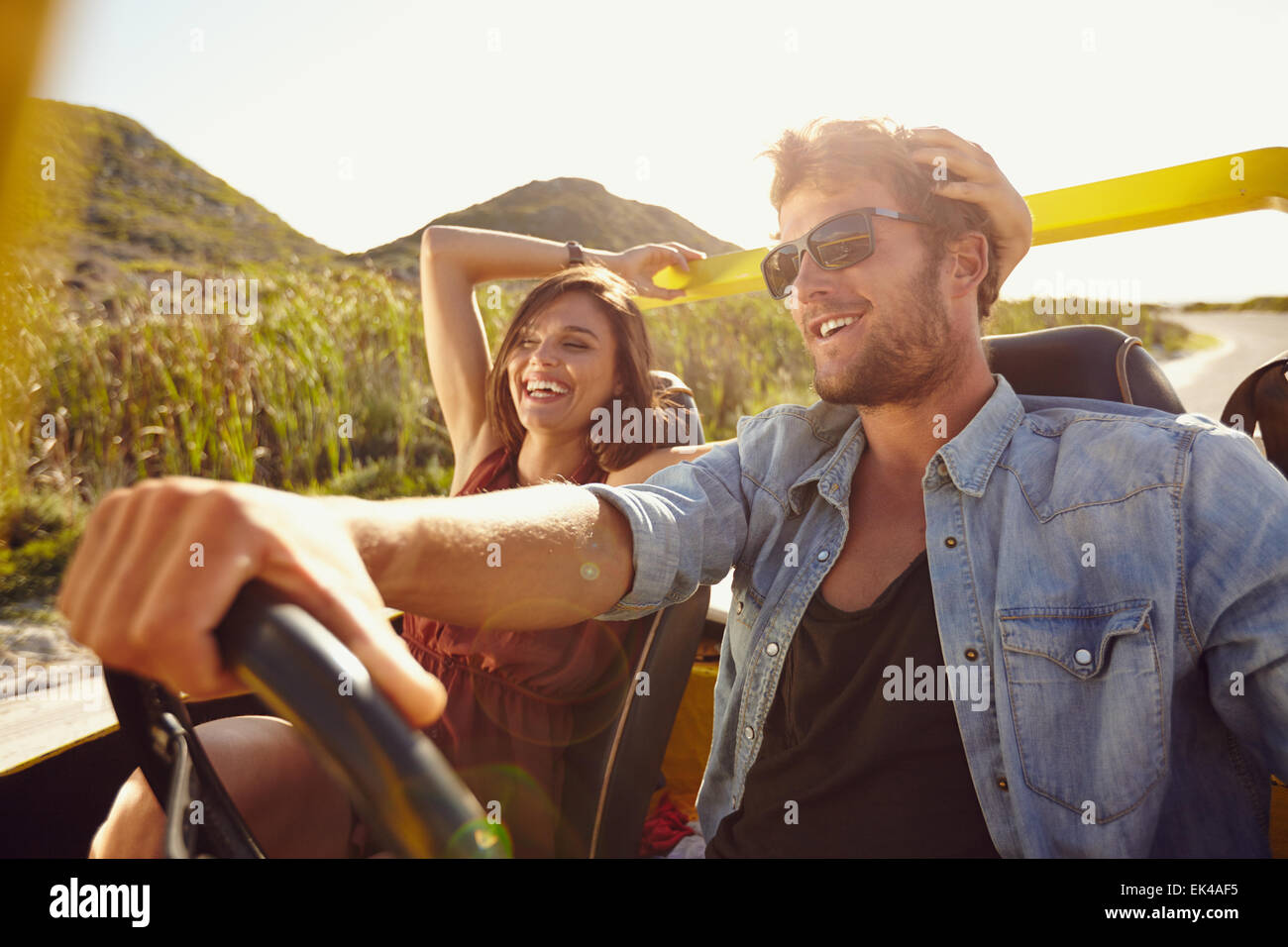 Cheerful young couple on road trip. Young man driving open topped car with woman smiling. Stock Photo