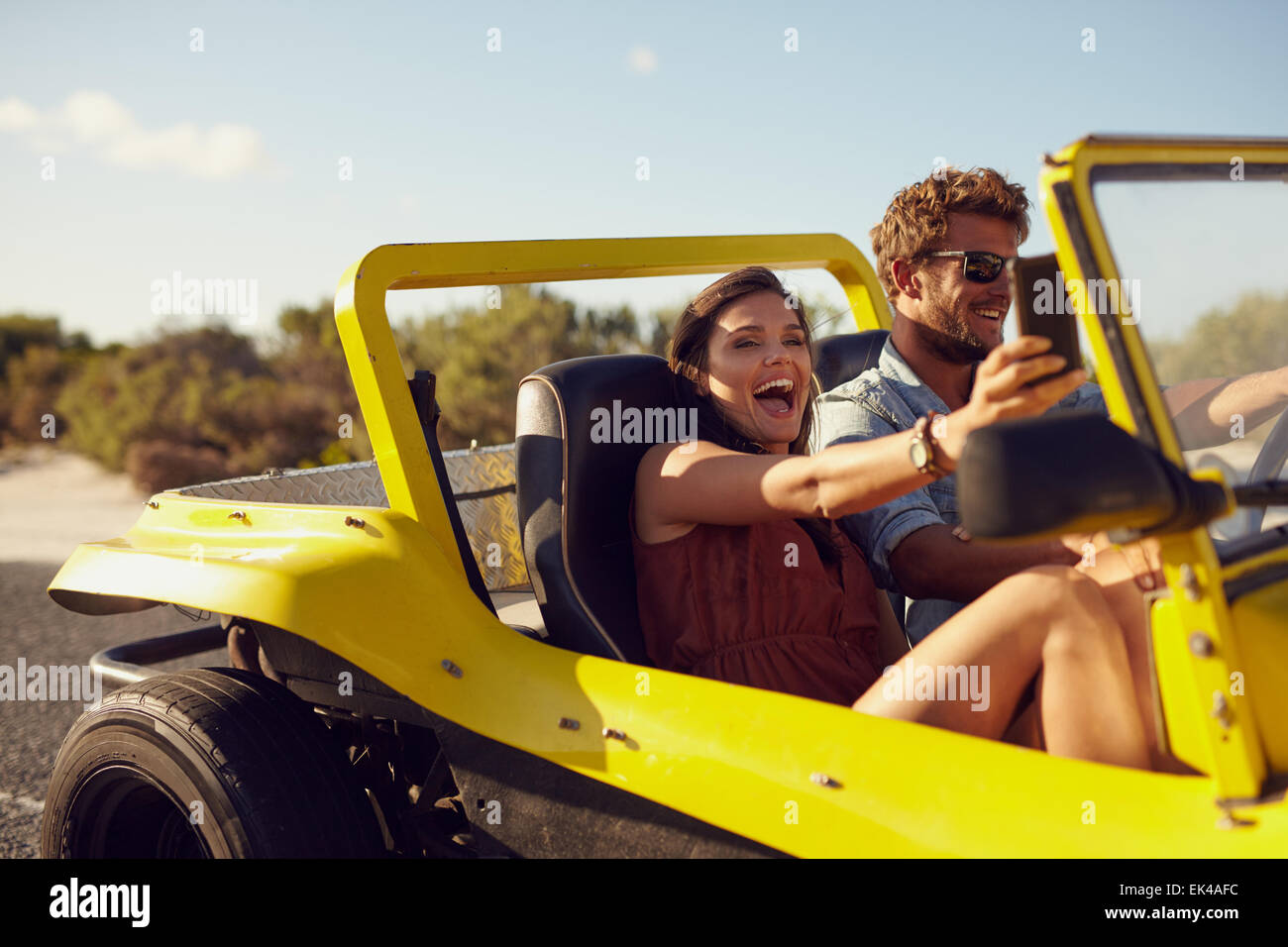 Excited happy couple enjoying road trip in their beach buggy. Man driving the car young woman taking a selfie from her mobile ph Stock Photo