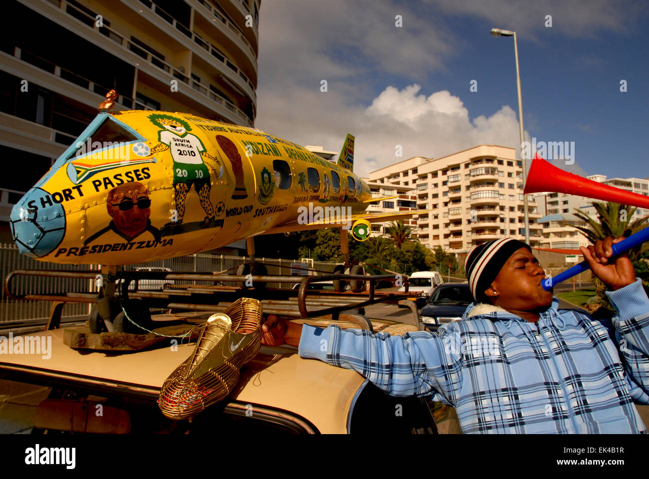 A man blows a vuvuzela next to  his car with a home made areoplane on the car to celebrebrate the 2010 FIFA World Cup in South Africa, Sea Point, Cape Town 09.06.2010. The aeroplane is made by Mboneli Lefty Booi and Siseko Christopher Kameso, both from Khayelitsha, a township in Cape Town.The plane is handmade and designed by them. It has been hand painted with a picture of Mandela, zakumi ( the south African world cup mascot), Zuma, Bafana Bafana etc It has all the flags painted on it of the countries taking part. They also made the boot. Their wish was,  “ we would like support from governme Stock Photo