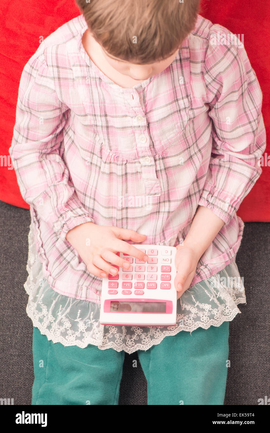 Little girl sitting on sofa at home with calculator in her hand. She is learning mathematics. Stock Photo