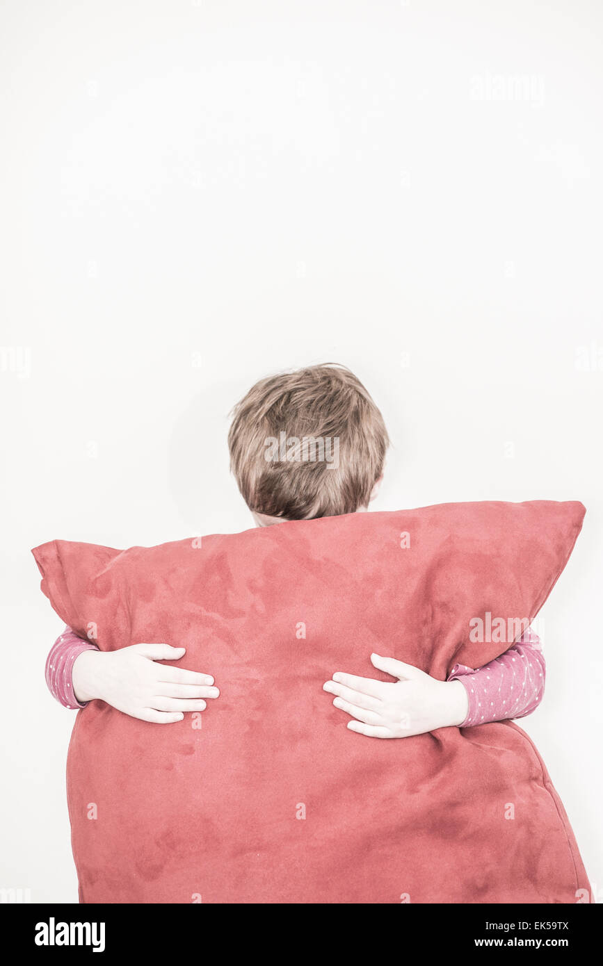 Sad little girl hiding behind a big red pillow. She is standing in