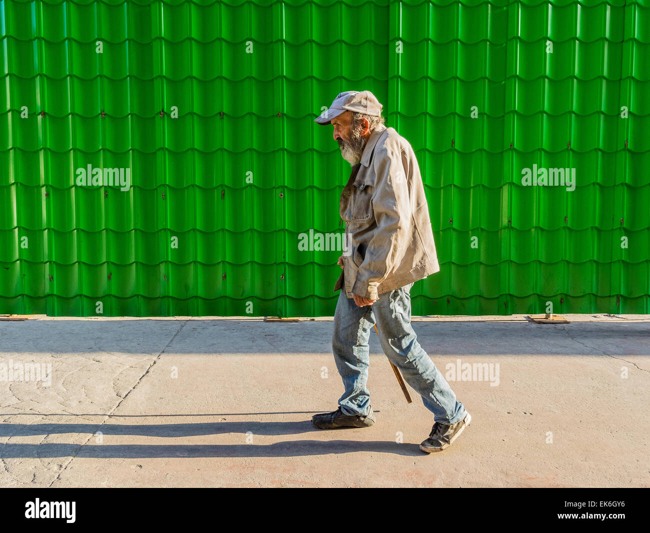 A male Hispanic senior citizen with a beard and wearing a baseball type hat, a soiled jacket and blue jeans walks with a cane be Stock Photo