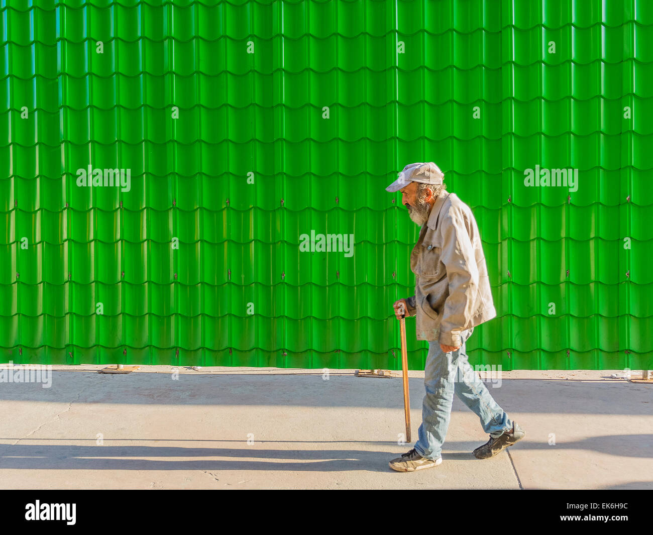 A male Hispanic senior citizen walks with a cane beside a bright green wall made of many tiles yielding a repetition of form. Stock Photo
