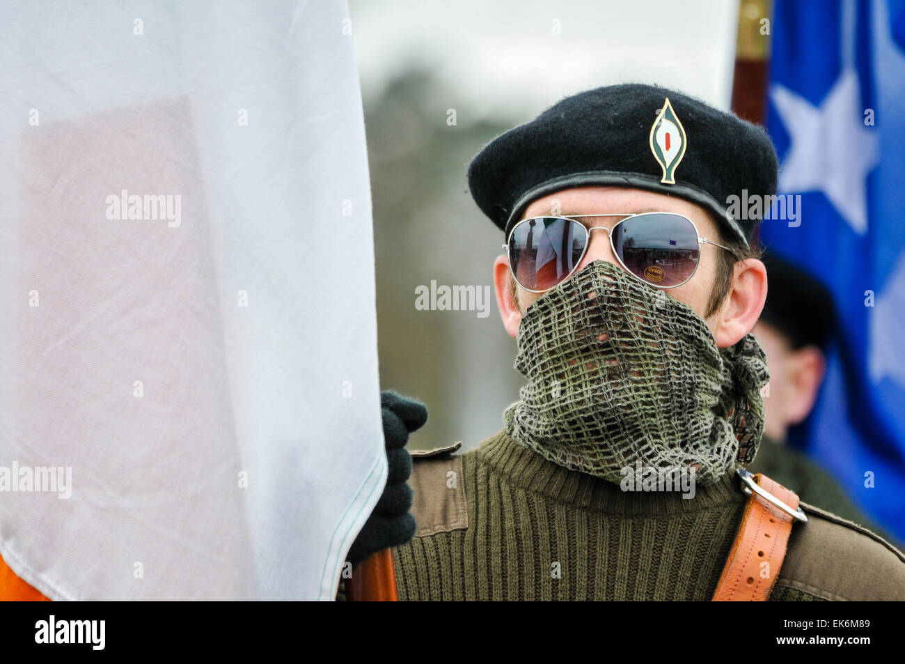 A masked man wearing a scrim over his mouth, sunglasses and a black beret holds a flag during an Irish Republican commemoration Stock Photo
