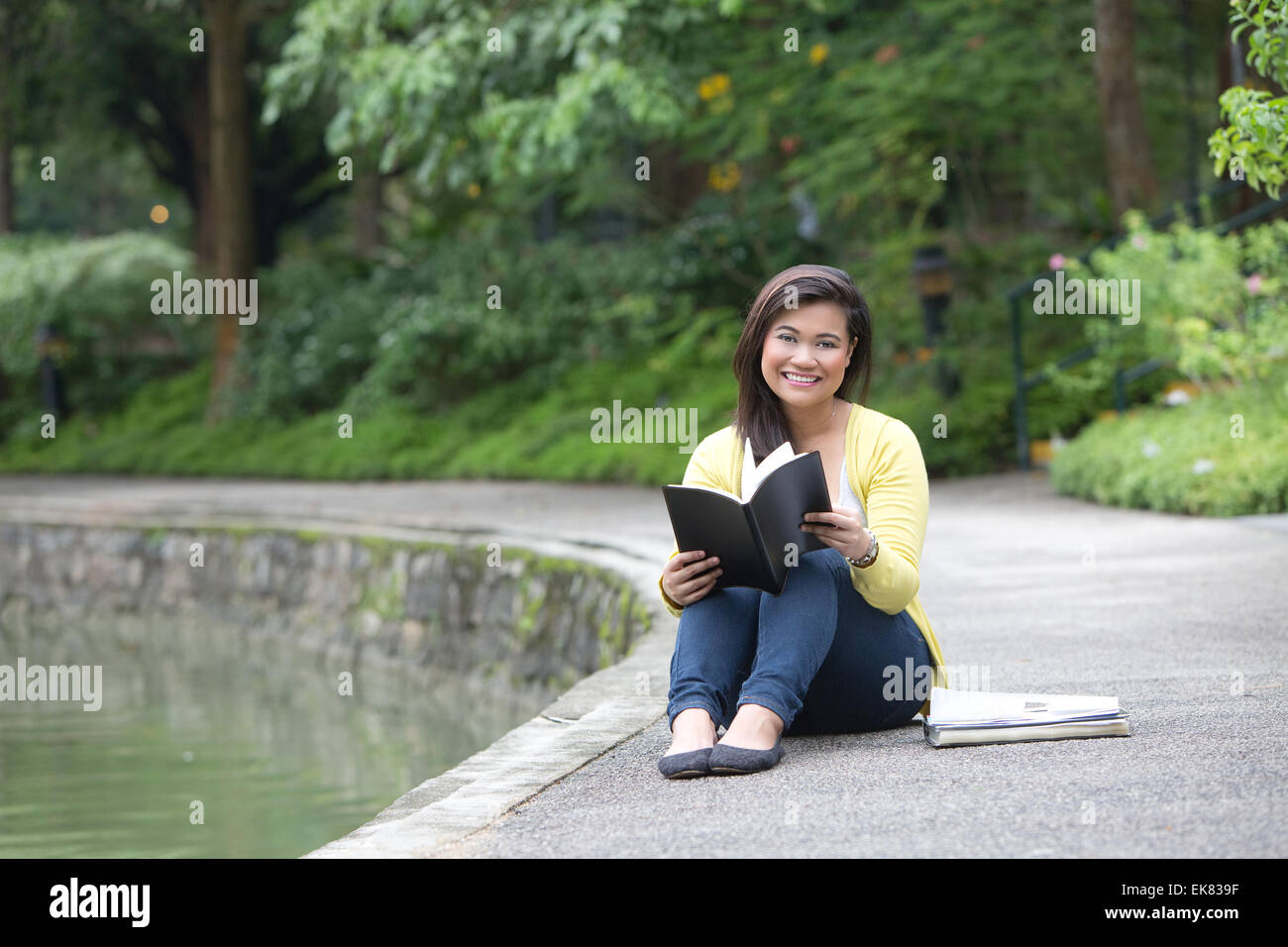 Beautiful young female university or college student smiling while holding a book, seated by a lake in a park. Stock Photo