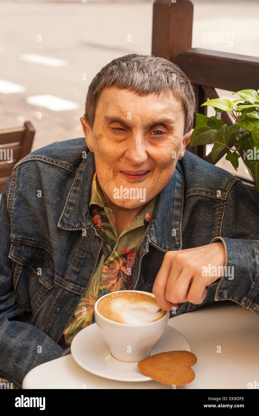 Elderly disabled man with cerebral palsy sitting at outdoor cafe with cup of coffee. Stock Photo