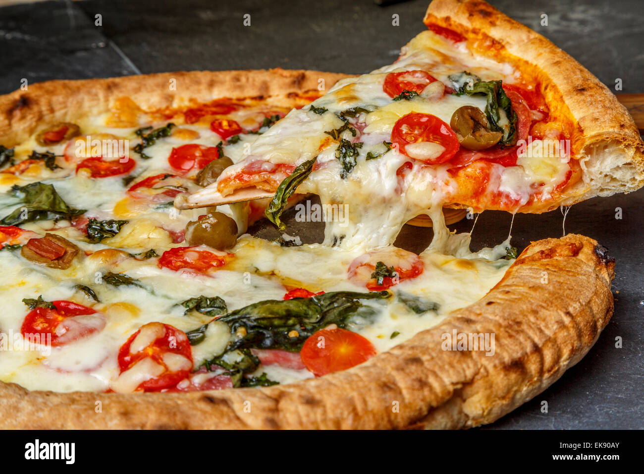 A close up view of a home made thin crust pizza slice with olives tomatoes peppers basil mozzarella cheese, London UK Stock Photo