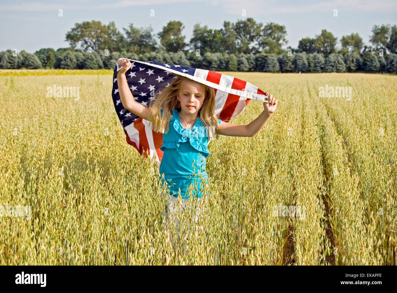 Young Caucasian girl running in a field of wheat with American flag. Stock Photo