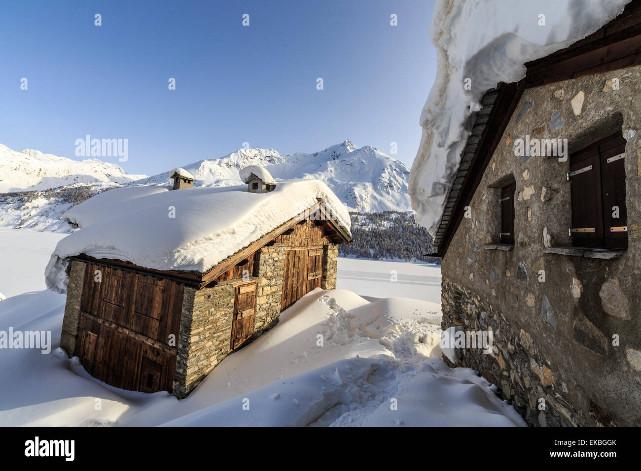 The sun, covered with thin clouds, illuminating a typical hut covered with snow, Graubunden, Swiss Alps, Switzerland Stock Photo
