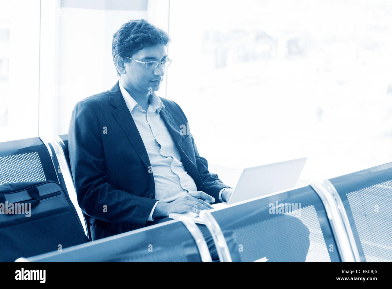 Asian Indian business man waiting his flight at airport, sitting on chair and using laptop, blue tone. Stock Photo