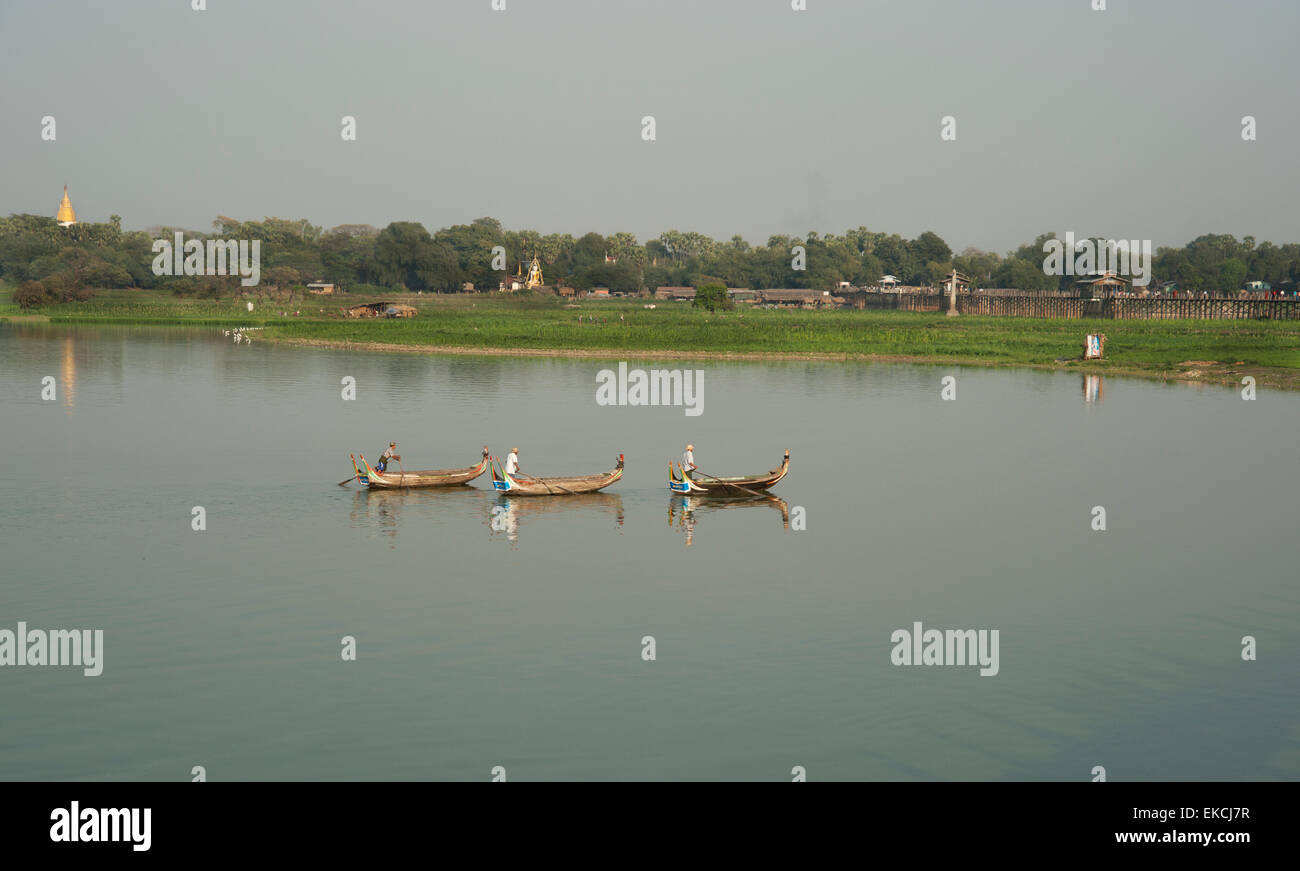 Three fishing boats glide on the placid waters of a lake in Myanmar on the shoreline people are working in the fields Stock Photo